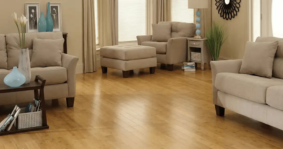 A living room with professionally installed bamboo flooring, beige sofas, a matching ottoman, a side table with a blue lamp, vases, framed pictures, and large windows with blinds and curtains in Dallas, TX